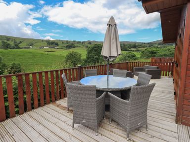 An outdoor wooden deck with a round glass table and six wicker chairs with a closed umbrella and two wicker armchairs overlooking green hills at Wansfell Retreat Lodge Wansfell 4 Limefitt Park near Windermere