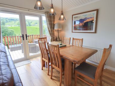 A dining area with a wooden table and six chairs next to glass doors leading to an outdoor deck with wicker furniture and green hillside view at Wansfell Retreat Lodge Wansfell 4 Limefitt Park near Windermere
