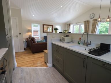 A kitchen with green cabinets and white countertop looking into a living room with a brown sofa and lamp at Wansfell Retreat Lodge Wansfell 4 Limefitt Park near Windermere