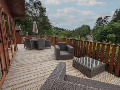 An outdoor wooden deck with wicker furniture including armchairs a glass coffee table a dining table with chairs and umbrellas at Wansfell Retreat Lodge Wansfell 4 Limefitt Park near Windermere
