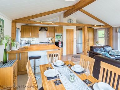 A dining area with a set table and an open kitchen with wooden cabinets and beams with a living area with leather sofas at Sheffield Pike Lodge Ullswater 8 Limefitt Park near Windermere