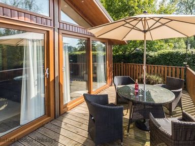 A wooden patio with a glass table and four wicker chairs under a large umbrella at Sheffield Pike Lodge Ullswater 8 Limefitt Park near Windermere
