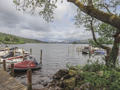 A lakeside view with boats docked at wooden piers and trees along the shore at Shoreside Lodge L8 Fallbarrow Park near Bowness on Windermere