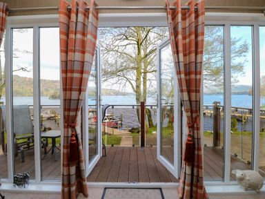 A room with open glass doors leading to a wooden deck overlooking a lake with trees and docks at Shoreside Lodge in L8 Fallbarrow Park near Bowness on Windermere