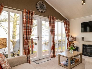 A living room with sliding glass doors and orange plaid curtains next to a wooden table with a vase of flowers at Shoreside Lodge in L8 Fallbarrow Park near Bowness on Windermere