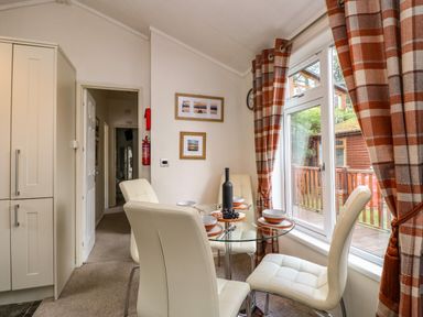 A dining area with a glass table set for four beside a window with red and beige check curtains at Shoreside Lodge L8 Fallbarrow Park near Bowness on Windermere