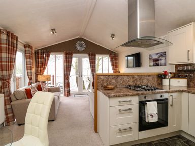 A kitchen with a gas stove and oven next to a living area with sofas and tartan curtains at Shoreside Lodge L8 Fallbarrow Park near Bowness on Windermere