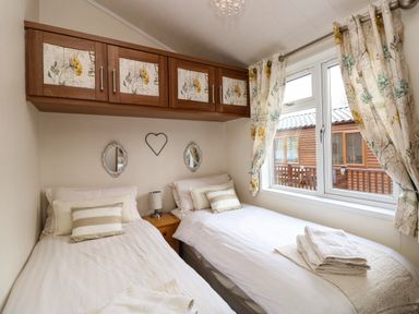 A bedroom with two single beds a window with curtains and overhead cabinets at Shoreside Lodge L8 Fallbarrow Park near Bowness on Windermere