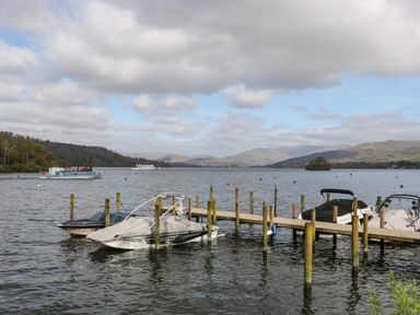 A lake with boats docked at a wooden pier and hills in the background at Shoreside Lodge L8 Fallbarrow Park near Bowness on Windermere