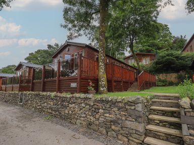 A wooden lodge with glass and wood railing on a raised stone platform with stairs and trees in the background at Shoreside Lodge L8 Fallbarrow Park near Bowness on Windermere