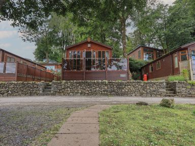 A wooden lodge with glass doors and a stone wall in front surrounded by trees at Shoreside Lodge L8 Fallbarrow Park near Bowness on Windermere