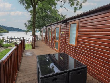 A wooden deck with railing outside a cabin by the water with a table and chairs at Shoreside Lodge L8 Fallbarrow Park near Bowness on Windermere