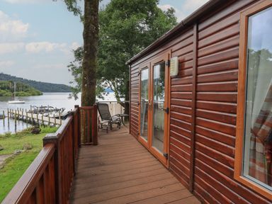 A wooden deck with chairs outside a cabin by the water at Shoreside Lodge L8 Fallbarrow Park near Bowness on Windermere