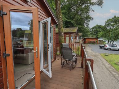 A wooden balcony with chairs outside wooden lodges near a parking area with cars and trees at Shoreside Lodge in L8 Fallbarrow Park near Bowness on Windermere