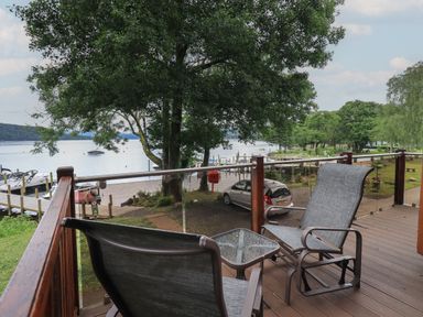 A balcony with two chairs and a glass table overlooking a lake with boats and trees at Shoreside Lodge L8 Fallbarrow Park near Bowness on Windermere