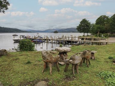 A lakeside area with wooden benches and table on grass near boats docked at a pier with hills in the background at Shoreside Lodge in L8 Fallbarrow Park near Bowness on Windermere