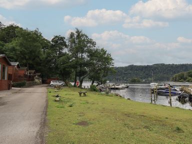 A lakeside view with wooden cabins on the left cars on a road trees near the water and boats docked at piers at Shoreside Lodge L8 Fallbarrow Park near Bowness on Windermere