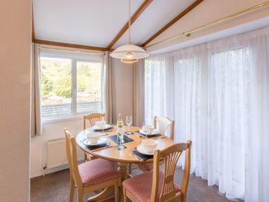 A dining area with a round wooden table set for four next to windows with curtains at Walkers Retreat Lodge Fellside 5 Limefitt Park near Windermere