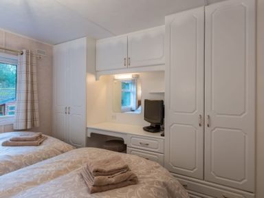 A bedroom with two beds with towels on them a window with curtains a built in wardrobe and a desk with a small television at Walkers Retreat Lodge Fellside 5 Limefitt Park near Windermere