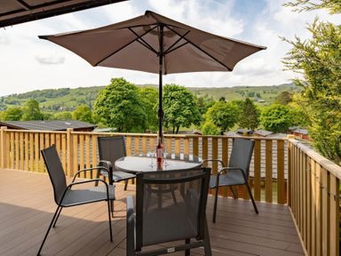 An outdoor wooden deck with a round glass table four chairs and a large umbrella overlooking trees and hills at Walkers Retreat Lodge Fellside 5 Limefitt Park near Windermere