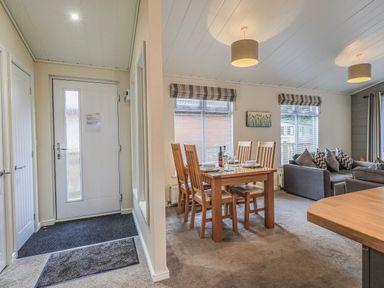 An entrance area with a white door leading to a dining area with a wooden table set for four and a living room with gray sofas at Daffodils Lodge Oakside 7 Fallbarrow Park in Bowness on Windermere