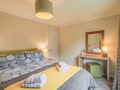 A bedroom with a bed with patterned bedding and folded towels a dressing table with a mirror a lamp and a green stool at Daffodils Lodge Oakside 7 Fallbarrow Park in Bowness on Windermere