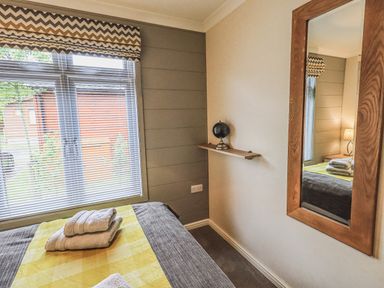 A bedroom with a bed, folded towels, a window with blinds, a shelf with a globe, and a wall mirror at Daffodils Lodge Oakside 7, Fallbarrow Park in Bowness on Windermere