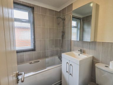 A bathroom with a bathtub shower, a sink with cabinet, a mirror, a window with blinds and a toilet at Daffodils Lodge Oakside 7 Fallbarrow Park in Bowness on Windermere