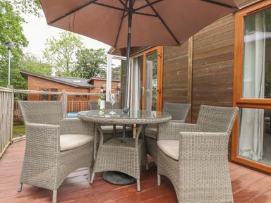 An outdoor wooden deck with wicker chairs around a glass table under a large umbrella at Daffodils Lodge Oakside 7 Fallbarrow Park in Bowness on Windermere
