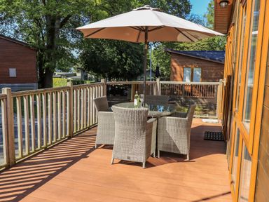 An outdoor deck with a round glass table seating four wicker chairs and a large umbrella at Daffodils Lodge in Oakside 7 Fallbarrow Park in Bowness on Windermere