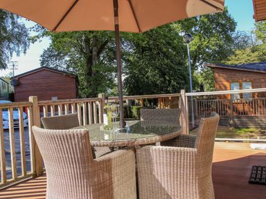 An outdoor deck with a round glass table, wicker chairs, an umbrella, and wooden cabins in the background at Daffodils Lodge Oakside 7 Fallbarrow Park in Bowness on Windermere