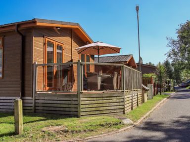 A wooden lodge with glass railing on the deck and outdoor table with umbrella at Daffodils Lodge Oakside 7 Fallbarrow Park in Bowness on Windermere