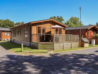 A wooden lodge with a fenced porch and umbrella beside a road at Daffodils Lodge Oakside 7 Fallbarrow Park in Bowness on Windermere