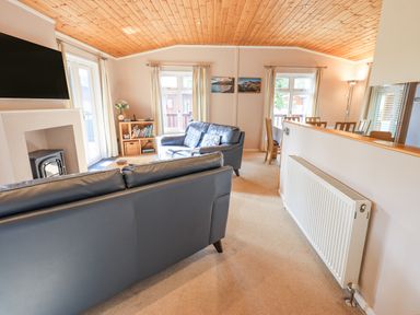 A living room with two black sofas a TV above a fireplace a bookshelf and a dining area at Tanglewood Lodge Thirlmere 15 White Cross Bay near Windermere