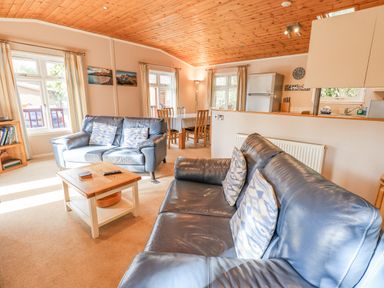 A living room with blue leather sofas a wooden coffee table a dining table with chairs and a kitchen area at Tanglewood Lodge in Thirlmere 15 White Cross Bay near Windermere