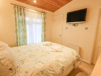A bedroom with a bed covered in floral bedding a wall-mounted television window with curtains and a wooden ceiling at Tanglewood Lodge in Thirlmere 15 White Cross Bay near Windermere