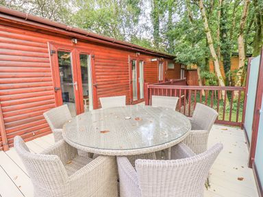 An outdoor deck area with a round glass table and six wicker chairs next to a wooden cabin at Tanglewood Lodge Thirlmere 15 White Cross Bay near Windermere