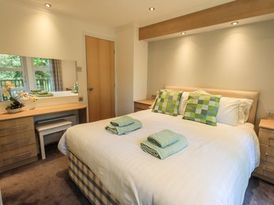 A bedroom with a double bed two green checkered pillows folded towels on bed bedside tables and a dressing table with a mirror at Forest Pines Lodge in Beech Hill 10 Fallbarrow Park near Bowness on Windermere