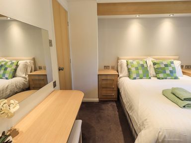 A bedroom with a bed two checkered pillows bedside tables and a wooden dressing table with a mirror at Forest Pines Lodge in Beech Hill 10 Fallbarrow Park near Bowness on Windermere