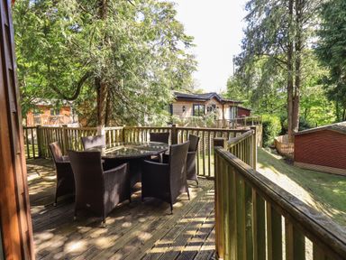 A wooden deck with a round glass table and six wicker chairs surrounded by trees at Forest Pines Lodge in Beech Hill 10 Fallbarrow Park near Bowness on Windermere