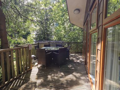 An outdoor wooden deck with a round glass table and six chairs surrounded by trees at Forest Pines Lodge Beech Hill 10 Fallbarrow Park near Bowness on Windermere