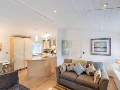 A living room with a sofa and cushions next to a kitchen area with a wooden breakfast bar at Broad Larch Lodge in Glade 16 Fallbarrow Park near Bowness on Windermere