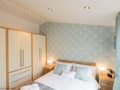 A bedroom with a bed and pillows two bedside tables with lamps and a wardrobe at Broad Larch Lodge in Glade 16 Fallbarrow Park near Bowness on Windermere