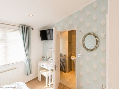 A bedroom with a window covered by curtains a wall-mounted television a vanity table with a stool and an open door leading to a bathroom at Broad Larch Lodge Glade 16 Fallbarrow Park near Bowness on Windermere