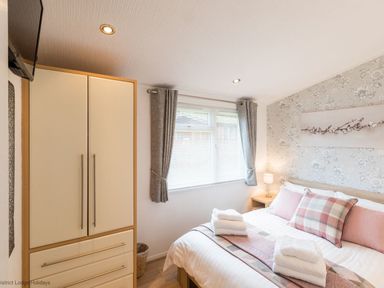 A bedroom with a bed, pillows, bedside table, lamp, wardrobe, window with curtains and floral wallpaper at Broad Larch Lodge in Glade 16 Fallbarrow Park near Bowness on Windermere