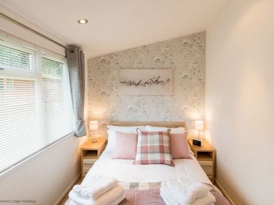 A bedroom with a double bed between two bedside tables with lamps and a window with blinds and curtains at Broad Larch Lodge in Glade 16 Fallbarrow Park near Bowness on Windermere