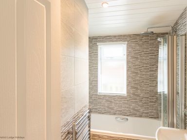 A bathroom with a bathtub shower combination a window with blinds a heated towel rail and tiled walls at Broad Larch Lodge in Glade 16 Fallbarrow Park near Bowness on Windermere