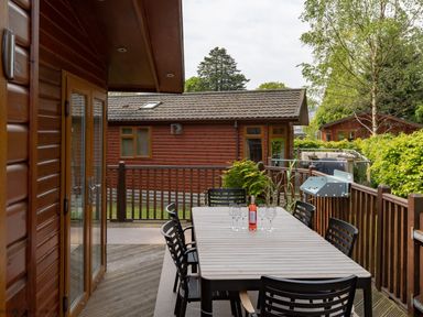An outdoor deck with a dining table and chairs with a bottle and glasses and wooden cabins in the background at Broad Larch Lodge in Glade 16 Fallbarrow Park near Bowness on Windermere