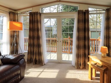 A living room with a leather armchair a floor lamp and a wooden side table with a lamp by glass doors with curtains at Squirrels Snug in Pony Meadow 1 White Cross Bay near Windermere
