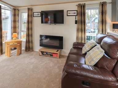 A living room with a brown leather sofa and two pillows a wall mounted television an electric fireplace a wooden side table with a lamp and patterned curtains at Squirrels Snug in Pony Meadow 1 White Cross Bay near Windermere